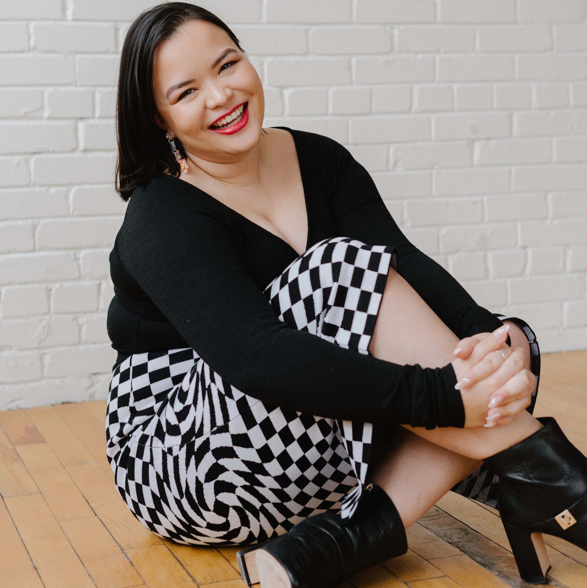 A smiling person sitting on a wooden floor, wearing a black long-sleeve top and a black-and-white checkered skirt, with black heels and a simple background of white brick walls.